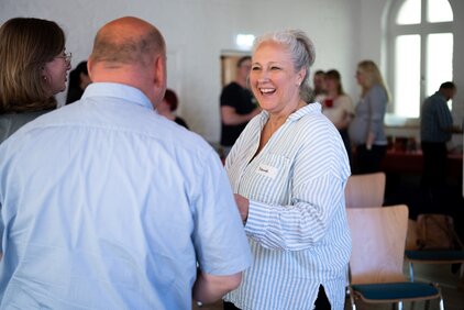 Eine Gruppe von Personen steht in einem Innenraum und unterhält sich. Eine Frau mit grauen Haaren und einem gestreiften Hemd lacht, während sie mit einem Mann spricht. Der Raum hat eine helllichte Atmosphäre und andere Personen sind im Hintergrund sichtbar.