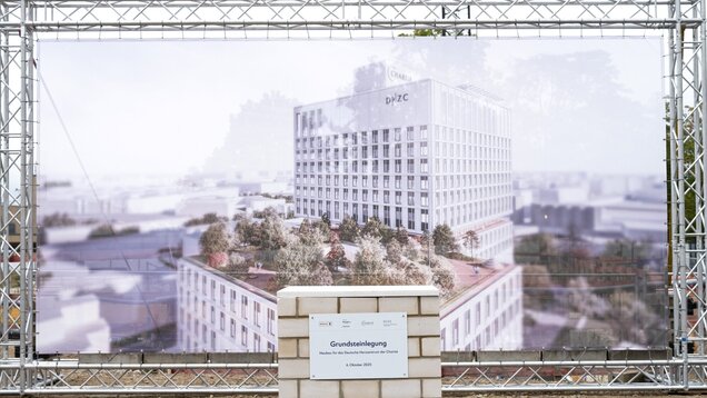 Ein Grundstein liegt auf einer Baustelle, vor einem großen Bauplan für das Deutsche Herzzentrum der Charité. Der Plan zeigt ein modernes Gebäude umgeben von Bäumen und Wegen. Das Datum der Grundsteinlegung ist der 6. Oktober 2023, dargestellt auf einer Tafel am Grundstein.