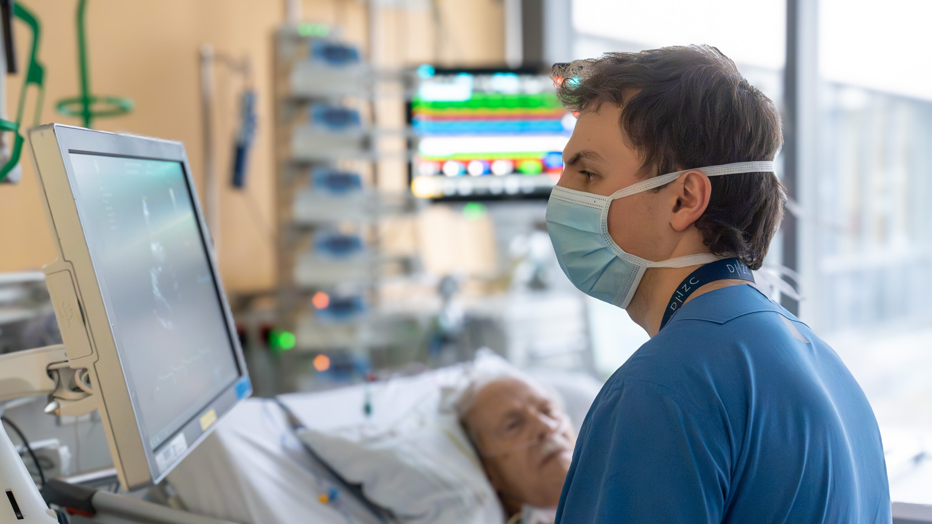 Ein Arzt in blauer Kleidung und chirurgischer Maske überwacht an einem Bildschirm die Vitaldaten eines Patienten im Krankenhausbett. Der Patient liegt bewusstlos da und wird medizinisch betreut.