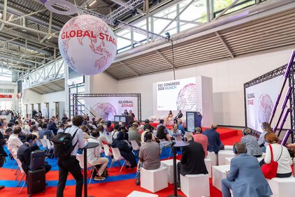 Eine Konferenzszene mit vielen Teilnehmern, die auf Stühlen sitzen und einem Podium mit der Aufschrift "GLOBAL STAGE" zusehen. Im Hintergrund sind große Bildschirme und ein globusförmiger Ball, der an der Decke hängt. Die Atmosphäre ist geschäftig und informativ.