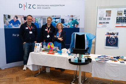 Drei Personen stehen an einem Tisch, der mit Informationsmaterialien und Werbeartikeln des Deutschen Herzzentrums der Charité bedeckt ist. Im Hintergrund ist ein Banner mit dem Logo des Zentrums zu sehen. Die Personen tragen blaue Sweatshirts und lächeln freundlich in die Kamera.