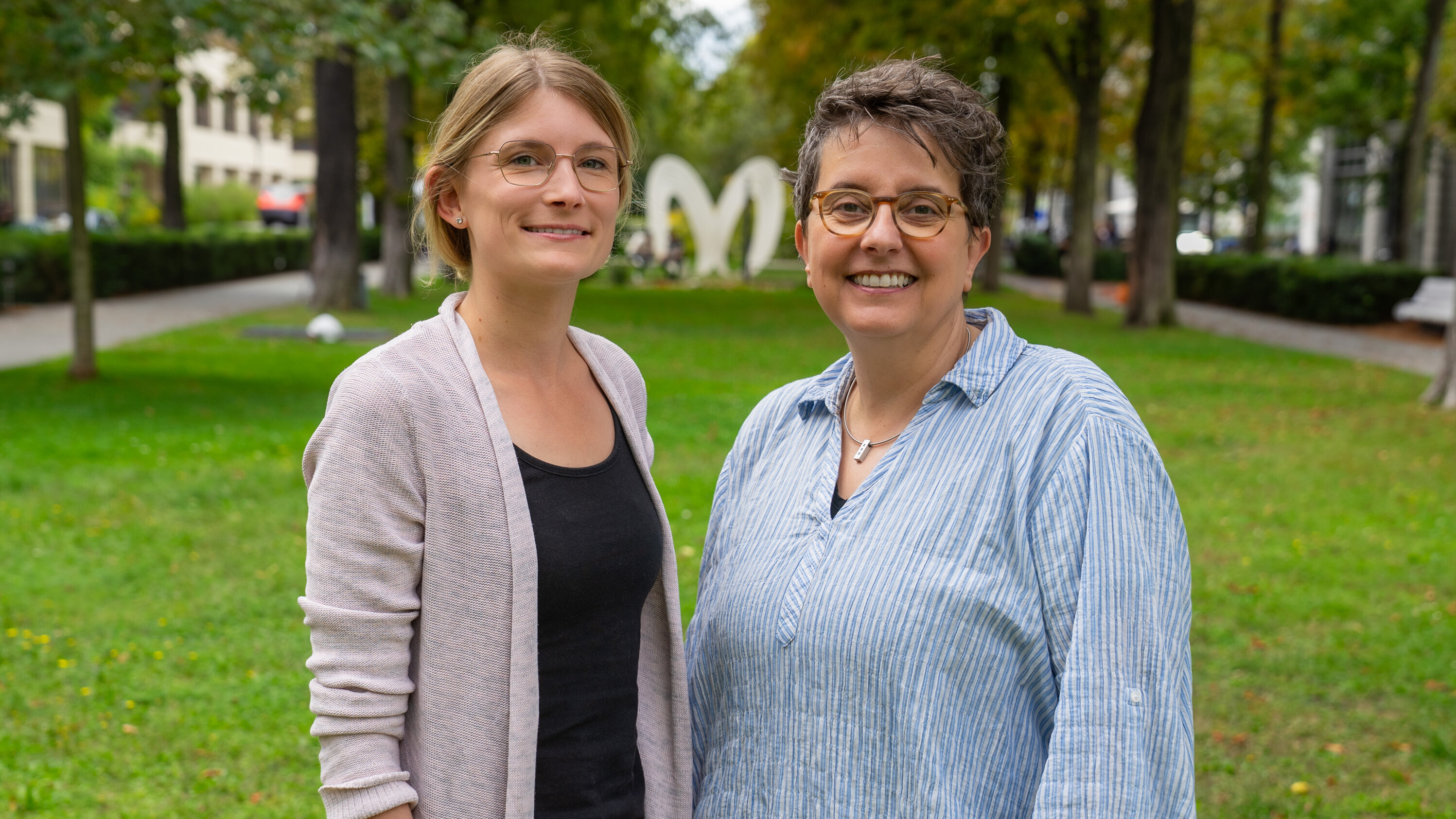 Zwei Frauen stehen lächelnd im Freien auf einer Wiese. Im Hintergrund sind Bäume und eine große, weiße Herzskulptur sichtbar. Die Frau links trägt ein helles Oberteil und eine Strickjacke, die Frau rechts ein gestreiftes Hemd.