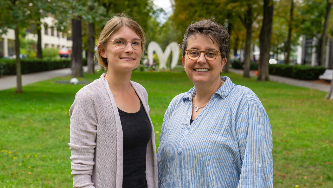 Zwei Frauen stehen lächelnd im Freien auf einer Wiese. Im Hintergrund sind Bäume und eine große, weiße Herzskulptur sichtbar. Die Frau links trägt ein helles Oberteil und eine Strickjacke, die Frau rechts ein gestreiftes Hemd.