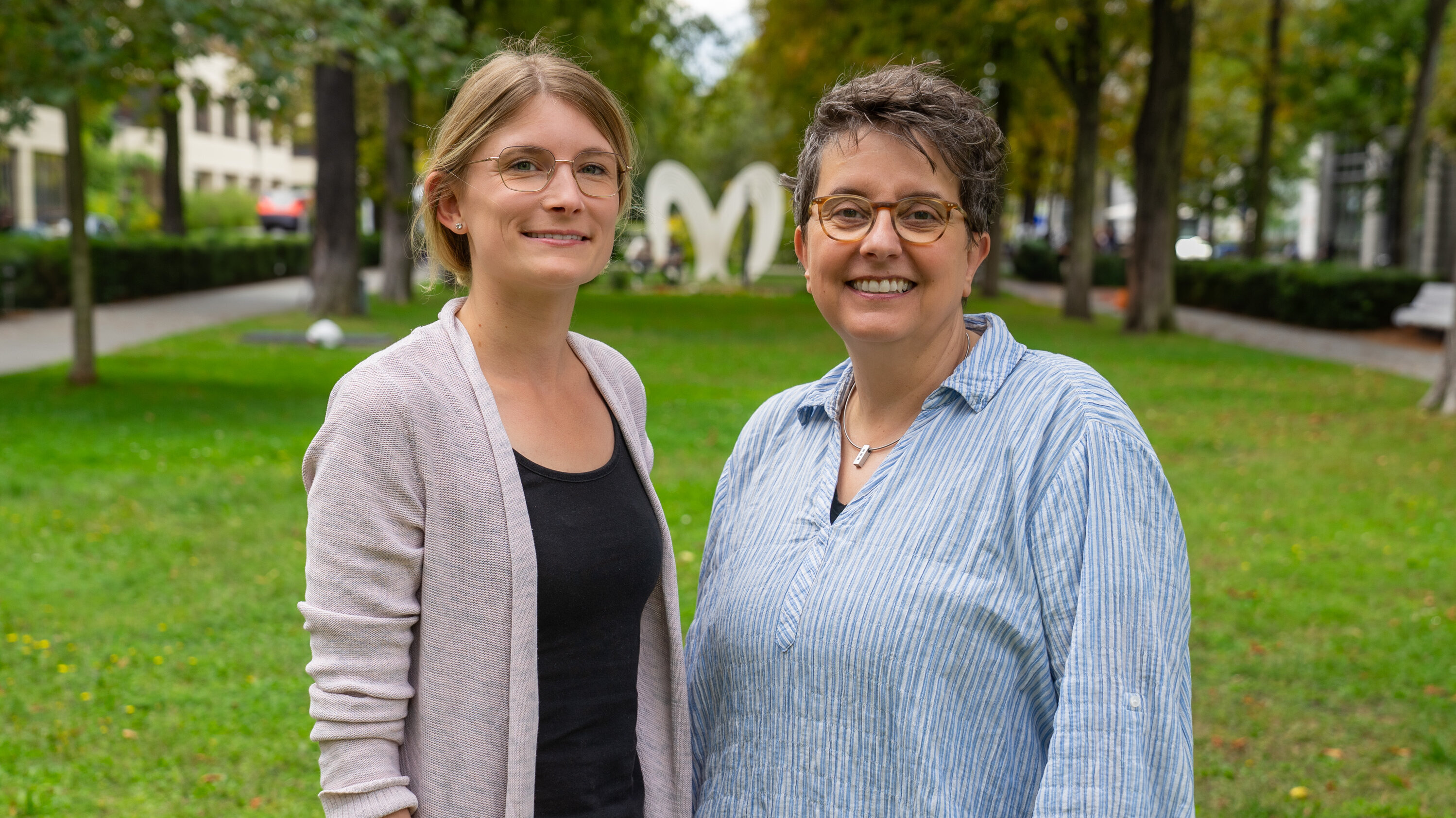 Zwei Frauen stehen lächelnd im Freien auf einer Wiese. Im Hintergrund sind Bäume und eine große, weiße Herzskulptur sichtbar. Die Frau links trägt ein helles Oberteil und eine Strickjacke, die Frau rechts ein gestreiftes Hemd.