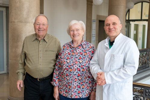 Ein älteres Paar und ein Arzt stehen lächelnd in einem hellen Innenraum. Der Mann trägt ein kariertes Hemd, die Frau ein blumiges Oberteil. Der Arzt in einem weißen Kittel hat die Hände vor sich verschränkt. Der Ausdruck auf ihren Gesichtern vermittelt eine freundliche Atmosphäre.
