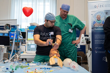 Zwei Personen in medizinischer Kleidung stehen an einem Tisch mit einem simulierten Patienten. Der eine Mann führt eine medizinische Prozedur durch, während der andere ihn beobachtet. Um sie herum sind verschiedene medizinische Geräte und Zubehörteile platziert. Im Hintergrund ist ein Herzballon zu sehen.