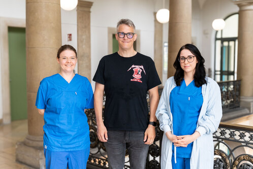 Drei Personen stehen in einem hellen Flurbereich. Zwei Frauen tragen medizinische Kleidung in Blau, während der Mann in einem schwarzen T-Shirt mit einem Aufdruck zu sehen ist. Sie lächeln und schauen in die Kamera. Der Hintergrund zeigt Säulen und eine moderne Architektur.