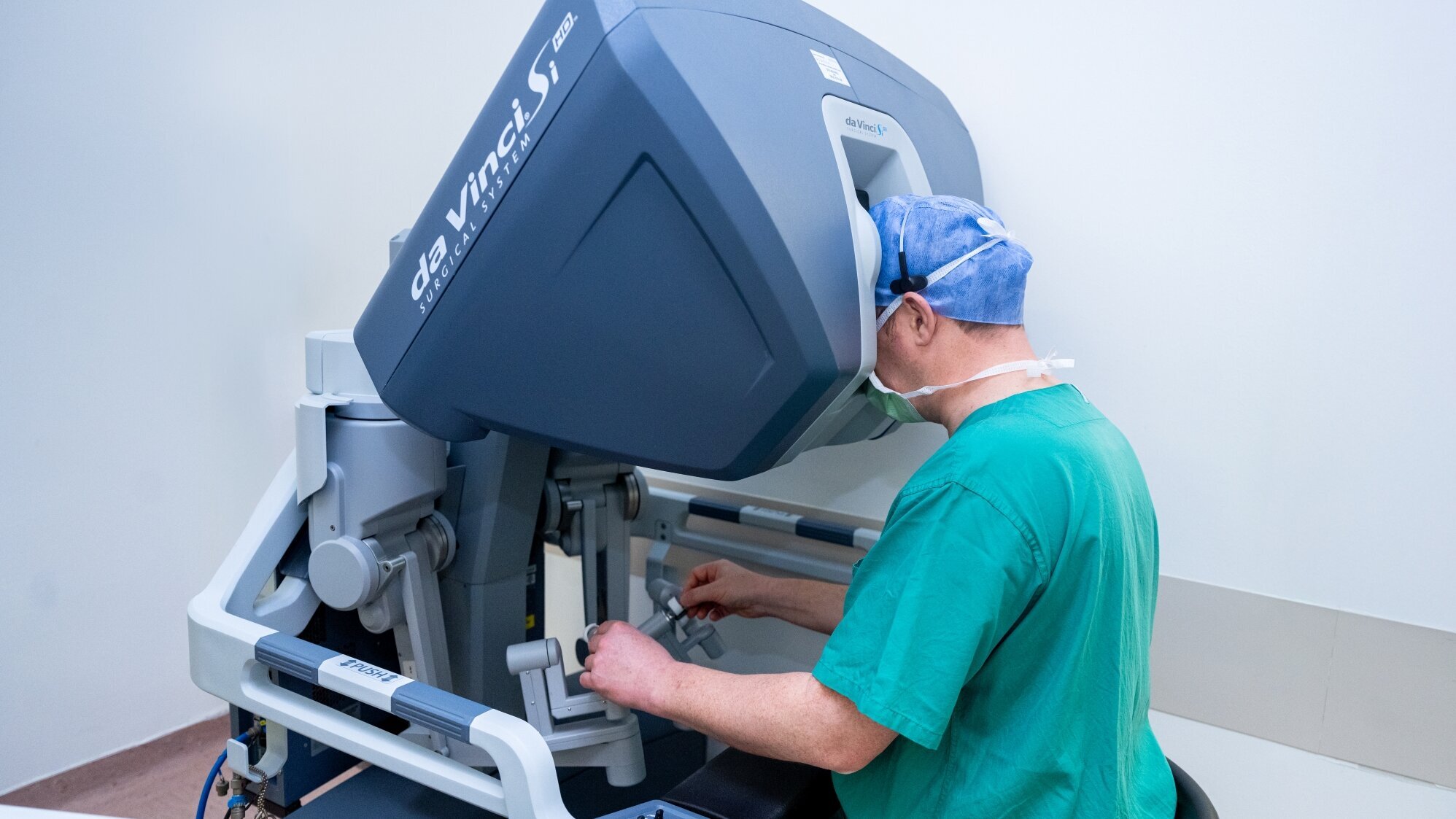 Ein Chirurg in grüner Kleidung bedient das da Vinci® Surgical System. Er sitzt an dem Roboter und konzentriert sich auf einen Bildschirm, während er die Steuerungselemente benutzt. Der Raum ist hell und minimalistisch gestaltet.