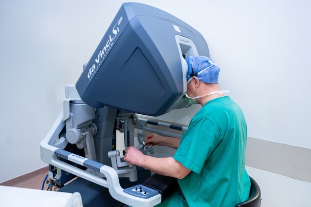 Ein Chirurg in grüner Kleidung bedient das da Vinci® Surgical System. Er sitzt an dem Roboter und konzentriert sich auf einen Bildschirm, während er die Steuerungselemente benutzt. Der Raum ist hell und minimalistisch gestaltet.