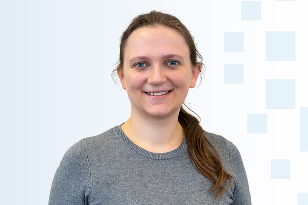 Porträt einer lächelnden Frau mit langen, braunen Haaren, die locker zu einer Seite gebunden sind. Sie trägt ein graues Sweatshirt und hat einen freundlichen Ausdruck. Der Hintergrund ist hell mit subtilen geometrischen Formen.