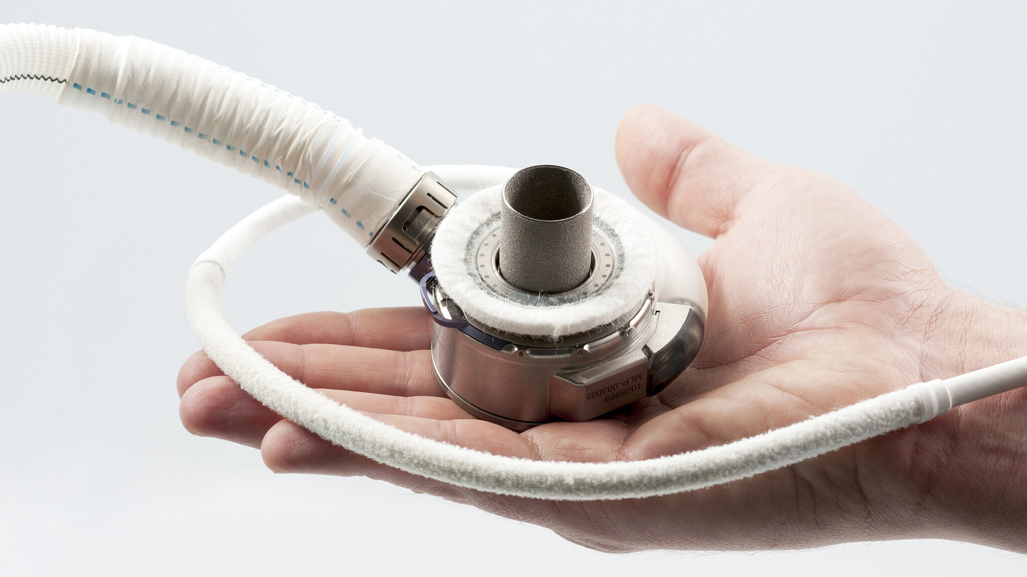 Eine Hand hält ein medizinisches Gerät, das aus einem runden, flachen Teil mit einem Metallrohr in der Mitte besteht. Um das Gerät verläuft ein flexibles, weißes Schlauchband. Der Hintergrund ist hell und schlicht.