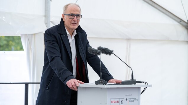 Ein Mann steht an einem Rednerpult, spricht in ein Mikrofon und wirkt konzentriert. Er trägt eine Brille und einen dunklen Mantel. Im Hintergrund sind Teile eines Zeltes sichtbar, das für eine Veranstaltung genutzt wird, möglicherweise in Berlin.