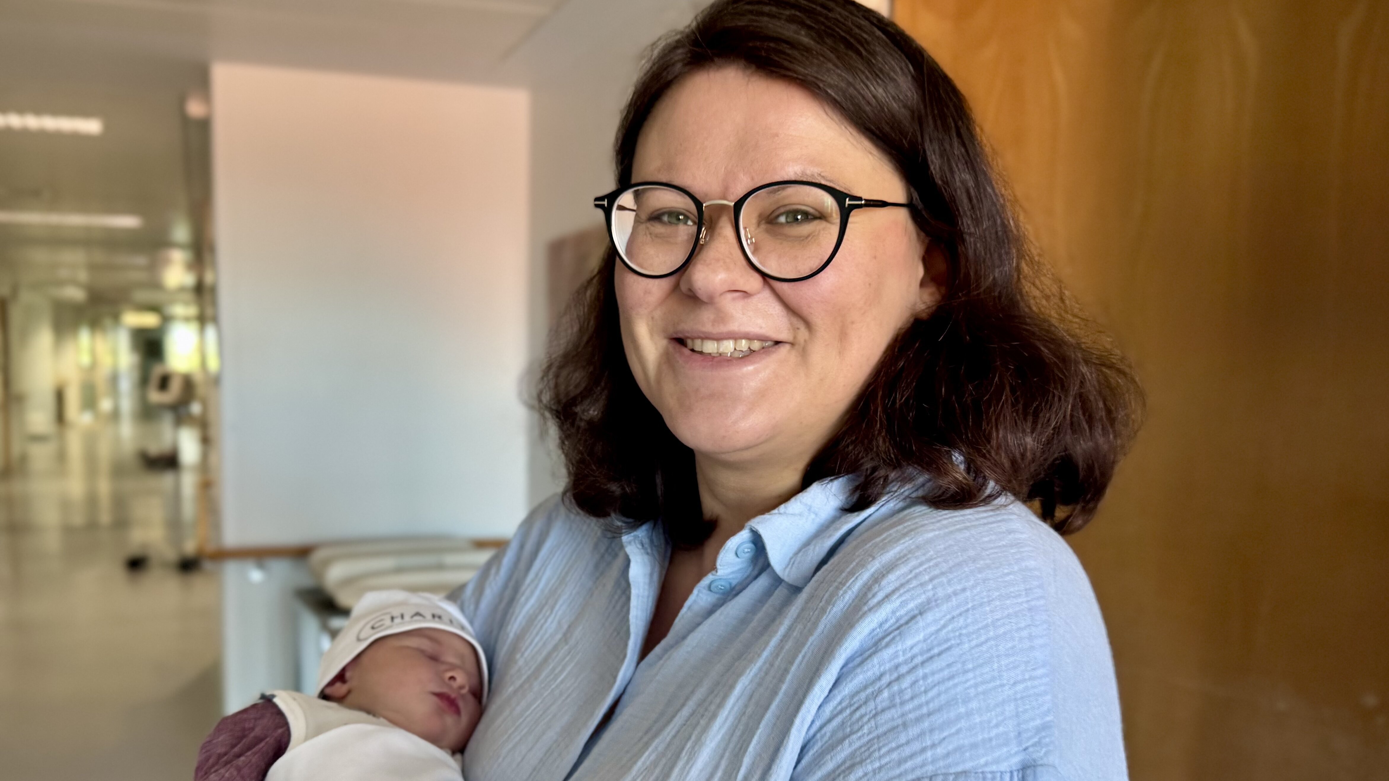 Eine lächelnde Frau mit Brille hält ein neugeborenes Baby in einem Krankenhausflur. Die Frau trägt eine hellblaue Bluse und hat lange, lockige Haare. Das Baby ist in eine weiße Decke gewickelt und trägt eine Mütze. Der Hintergrund zeigt einen modernen Krankenhausbereich.