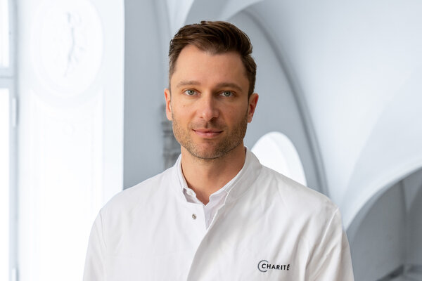 Ein Mann in einem weißen Arztkittel steht vor einem hellen, modernen Hintergrund. Er schaut direkt in die Kamera und vermittelt Professionalität und Freundlichkeit.