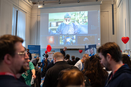 Eine Menschenmenge versammelt sich in einem Raum, während auf einer großen Leinwand eine Operation übertragen wird. Im Vordergrund sind mehrere Personen zu sehen, die interessiert auf die Präsentation schauen. Rote Herzen und Informationsstände sind ebenfalls im Raum vorhanden.