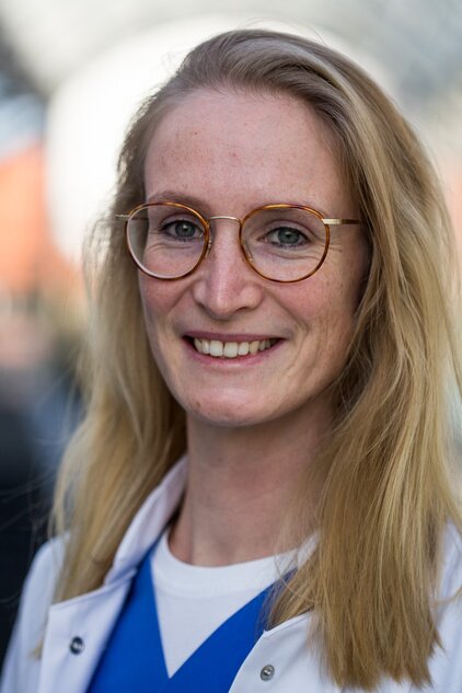 Ein junger Mensch mit langen, blonden Haaren und einer weißen Jacke lächelt freundlich in die Kamera. Sie trägt eine Brille mit runden, goldfarbigen Rahmen. Der Hintergrund ist unscharf und zeigt undeutliche Farben.
