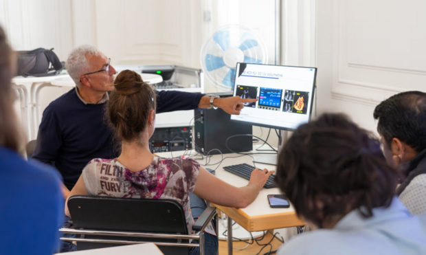 Ein Dozent erklärt in einem Seminarraum etwas zu 3D-Volumetrie und zeigt auf Inhalte auf einem Bildschirm. Eine Gruppe von Teilnehmern hört aufmerksam zu, während eine Person am Laptop arbeitet.