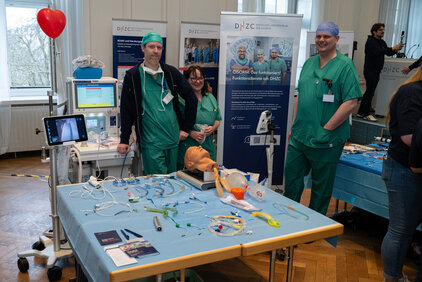 Eine Gruppe von medizinischem Personal in grüner Kleidung steht neben einem Tisch, der mit medizinischen Geräten und Instrumenten ausgestattet ist. Im Hintergrund sind informative Plakate über Anästhesie zu sehen. Die Atmosphäre wirkt professionell und informativ, passend für eine medizinische Veranstaltung.