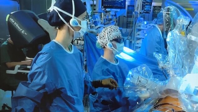 Das Bild zeigt mehrere Ärzte in blauer OP-Kleidung, die an einem Patienten arbeiten. Sie tragen Masken und spezielle Kopfbedeckungen. Im Hintergrund sind medizinische Geräte und Bildschirme zu sehen, die eine chirurgische Umgebung darstellen.