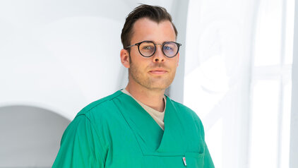 Ein junger Mann trägt einen grünen Arztkittel und eine Brille. Er blickt direkt in die Kamera und steht vor einem hellen, unscharfen Hintergrund. Sein Gesichtsausdruck ist neutral und konzentriert.