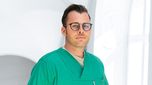 Ein junger Mann trägt einen grünen Arztkittel und eine Brille. Er blickt direkt in die Kamera und steht vor einem hellen, unscharfen Hintergrund. Sein Gesichtsausdruck ist neutral und konzentriert.