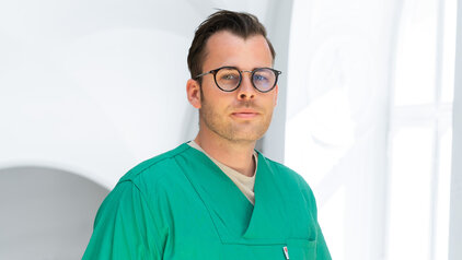 Ein junger Mann trägt einen grünen Arztkittel und eine Brille. Er blickt direkt in die Kamera und steht vor einem hellen, unscharfen Hintergrund. Sein Gesichtsausdruck ist neutral und konzentriert.