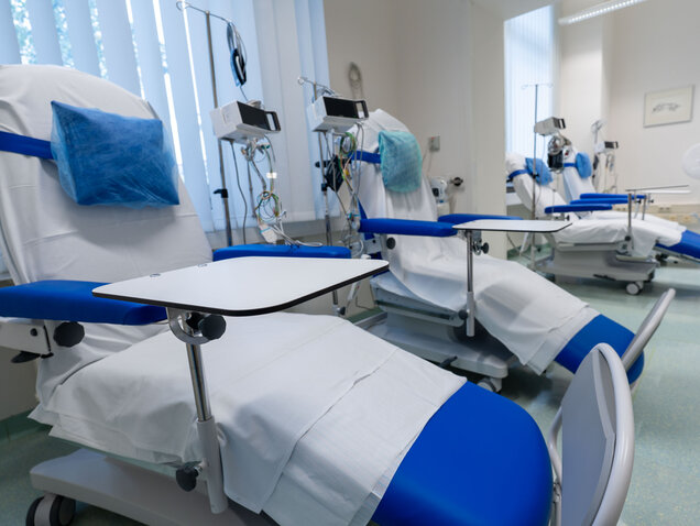 Ein Raum mit mehreren medizinischen Behandlungsstühlen, die mit weißen Laken und blauen Kissen ausgestattet sind. Jeder Stuhl hat einen kleinen Tisch und ist mit medizinischen Geräten verbunden. Der Raum wirkt sauber und ordentlich, mit viel natürlichem Licht.
