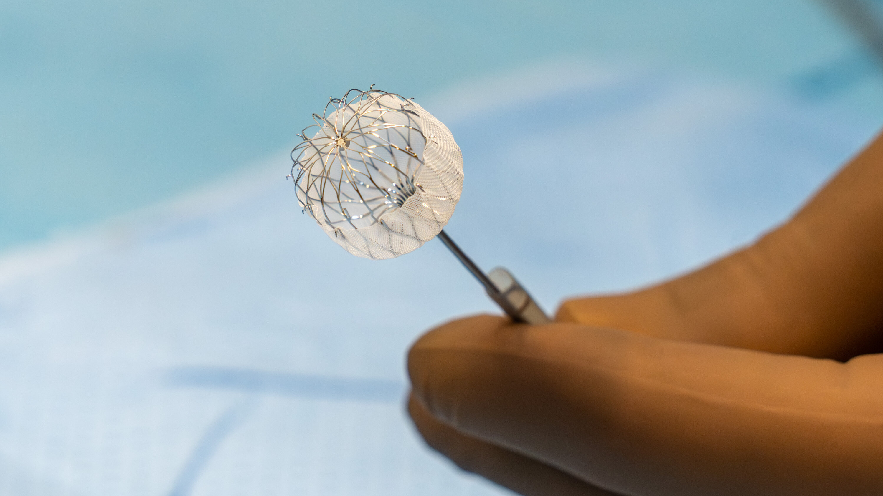 Eine Hand mit einem chirurgischen Instrument hält ein netzartiges, ballförmiges medizinisches Gerät. Der Hintergrund ist unscharf und zeigt eine sterile Umgebung, typisch für einen medizinischen Eingriff.