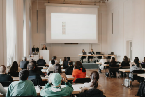 Eine Gruppe von Personen sitzt in einem Raum mit Tischen und hört einem Vortrag zu. An einer Leinwand sind Präsentationsinhalte zu sehen. Vorne steht ein Redner, während andere auf einer Panel-Bühne sitzen. Der Raum hat große Fenster und helle Wände.