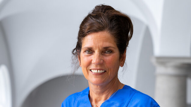 Eine lächelnde Frau in einem blauen Oberteil steht vor einem neutralen Hintergrund. Ihr Haar ist zu einem lockeren Dutt gesteckt. Sie wirkt freundlich und engagiert, bereit, ihre Arbeit zu tun.