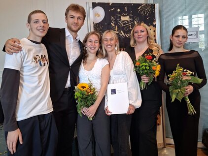 Sechs junge Erwachsene stehen in einer Gruppe. Sie halten Blumensträuße und eine Urkunde. Die Kleidung variiert von formell bis lässig. Die Szene strahlt Freude und Feierlichkeit aus, möglicherweise nach einer Abschlussverleihung oder ähnlichem Anlass. Licht strömt durch große Fenster im Hintergrund.