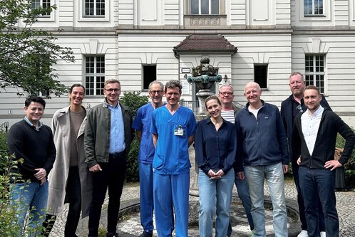 Eine Gruppe von zehn Personen steht vor einem Gebäude, das historischen Charme ausstrahlt. In der Mitte tragen einige Personen blaue Krankenhauskleidung. Alle lächeln und wirken freundlich. Im Hintergrund ist ein Brunnen und grüne Bepflanzung sichtbar.