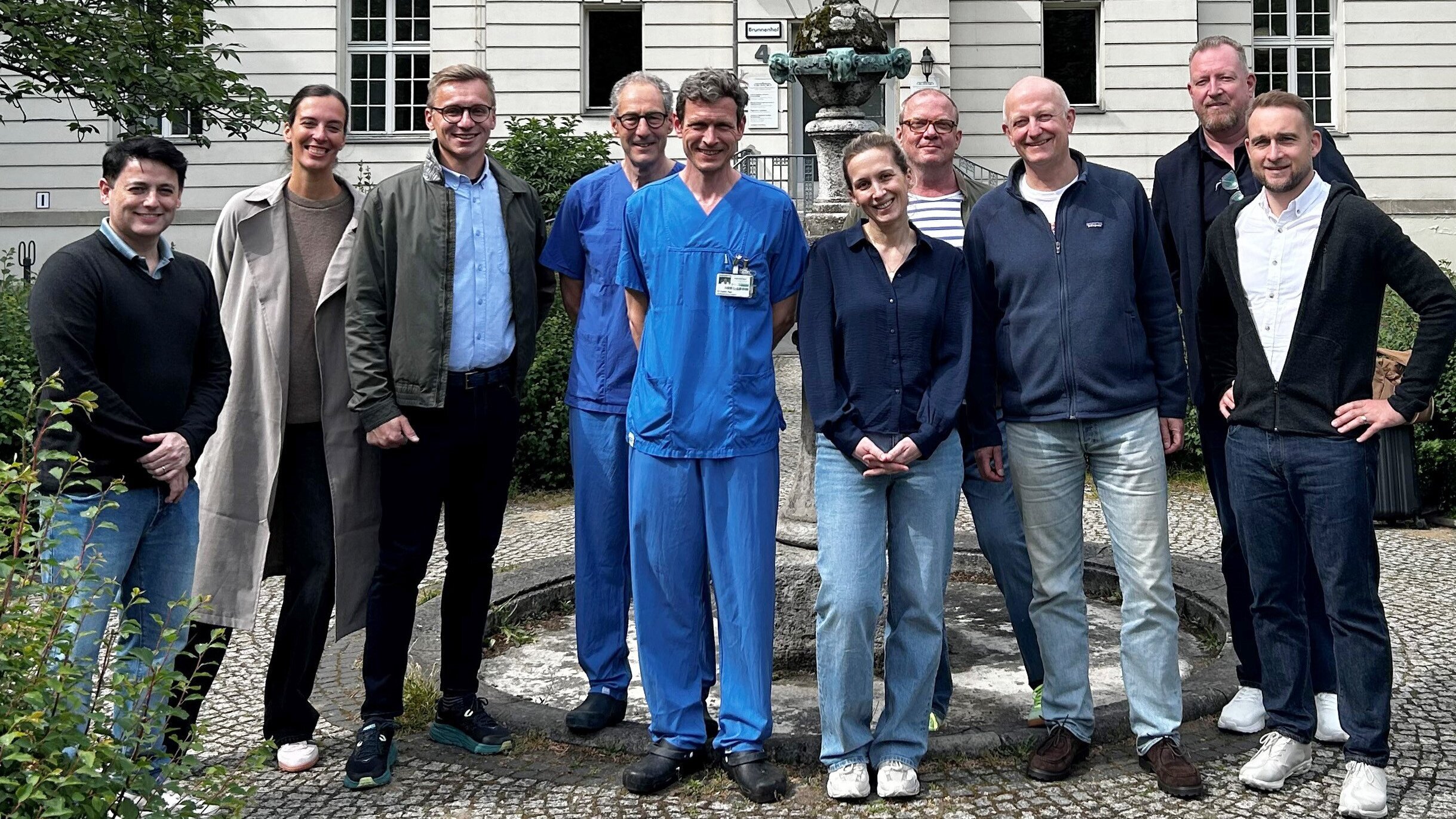 Eine Gruppe von zehn Personen steht vor einem Gebäude, das historischen Charme ausstrahlt. In der Mitte tragen einige Personen blaue Krankenhauskleidung. Alle lächeln und wirken freundlich. Im Hintergrund ist ein Brunnen und grüne Bepflanzung sichtbar.