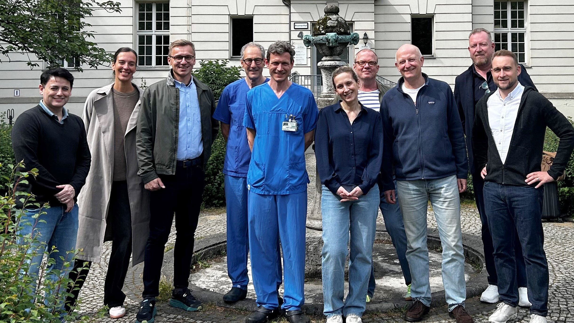 Eine Gruppe von zehn Personen steht vor einem Gebäude, das historischen Charme ausstrahlt. In der Mitte tragen einige Personen blaue Krankenhauskleidung. Alle lächeln und wirken freundlich. Im Hintergrund ist ein Brunnen und grüne Bepflanzung sichtbar.