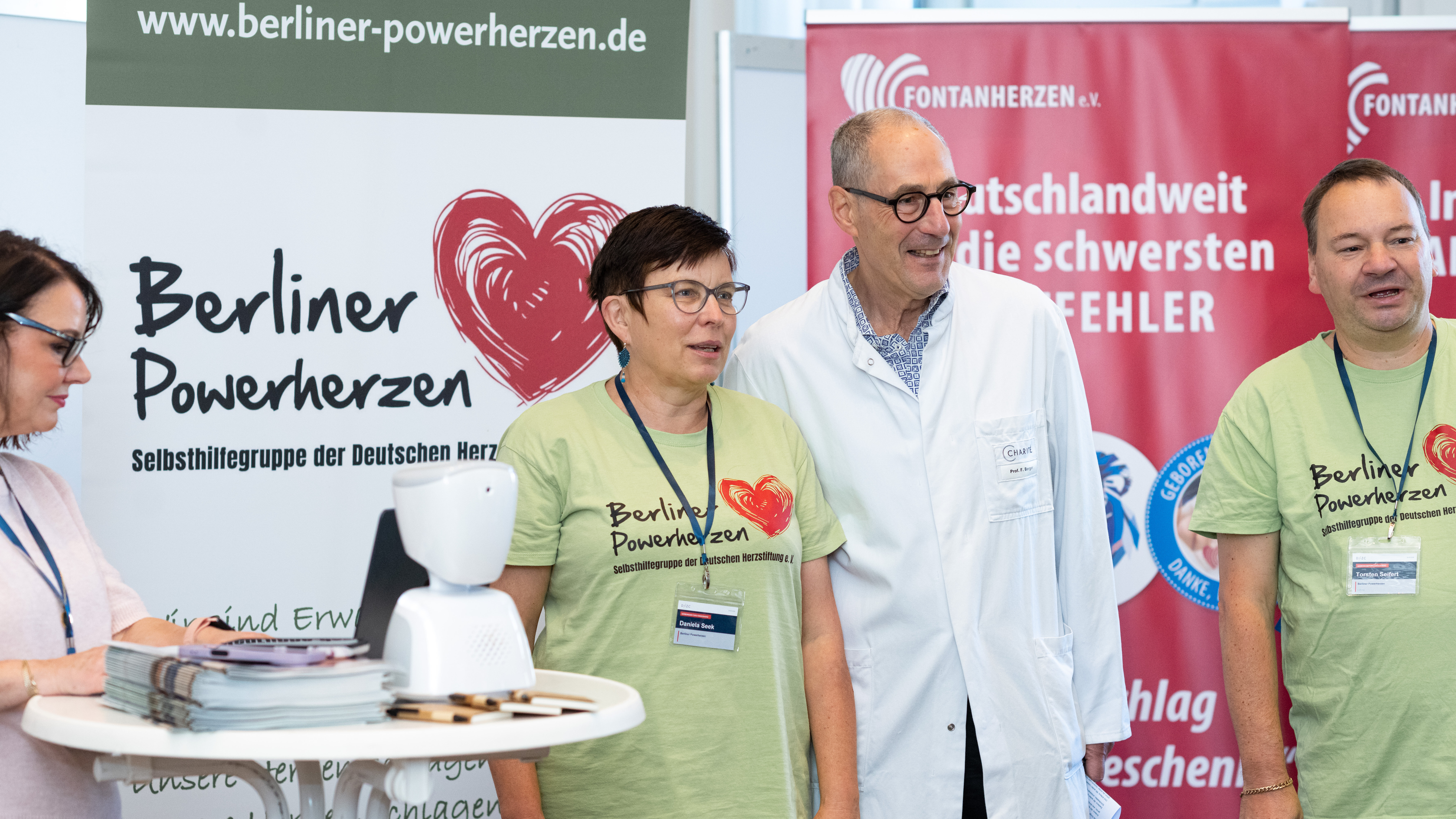 Eine Gruppe von Menschen steht zusammen in einem Raum. Zwei Frauen in grünen T-Shirts repräsentieren die Initiative „Berliner Powerherzen“. Ein Mann in einem weißen Kittel ist ebenfalls anwesend. Im Hintergrund sind Informationsbanner zu sehen, die sich mit Herzgesundheit beschäftigen.