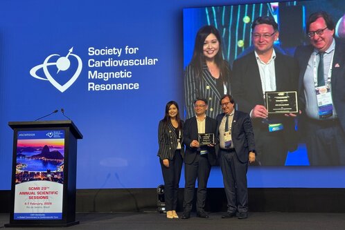 Auf der Bühne stehen vier Personen, die Anerkennungen in den Händen halten. Im Hintergrund ist das Logo der Gesellschaft für kardiovaskuläre Magnetresonanz zu sehen. Die Veranstaltung scheint sich um wissenschaftliche Themen im Bereich der Kardiologie zu drehen.