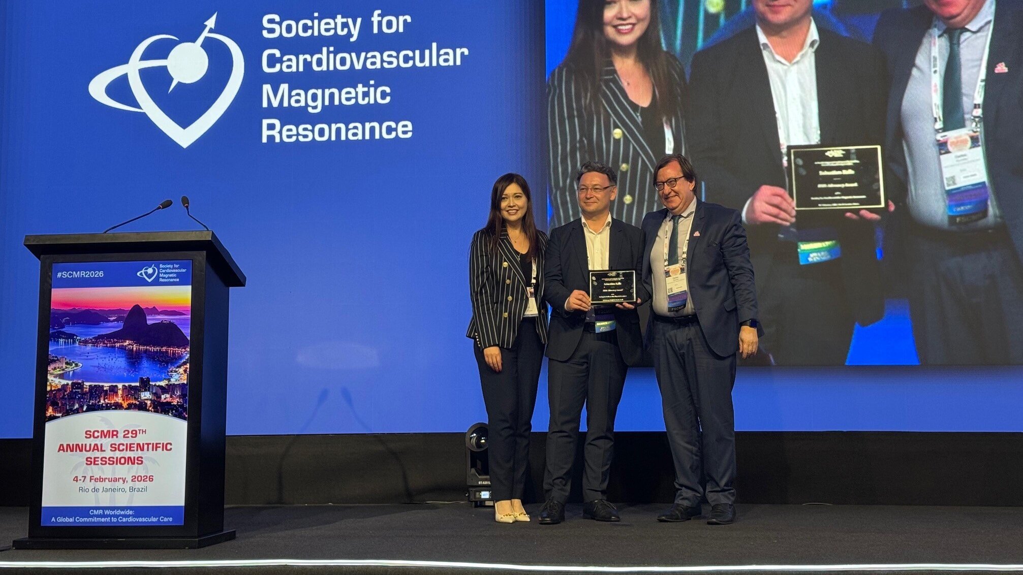 Auf der Bühne stehen vier Personen, die Anerkennungen in den Händen halten. Im Hintergrund ist das Logo der Gesellschaft für kardiovaskuläre Magnetresonanz zu sehen. Die Veranstaltung scheint sich um wissenschaftliche Themen im Bereich der Kardiologie zu drehen.