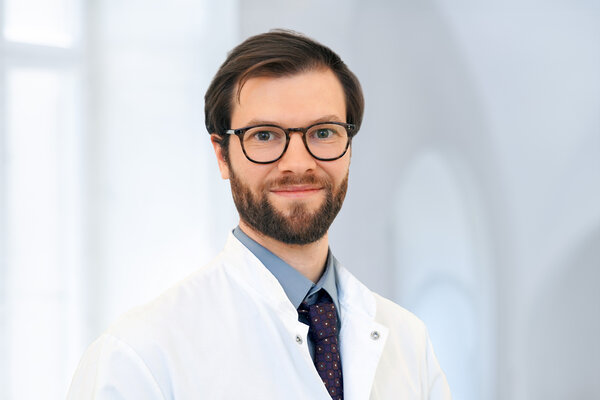 Ein junger Arzt mit Bart trägt eine weiße Arztjacke, ein hellblaues Hemd und eine dunkelblaue Krawatte. Er steht lächelnd vor einem hellen, unscharfen Hintergrund. Seine Brille hat einen schmalen Rahmen.