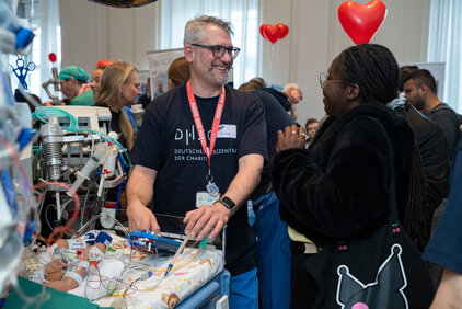 Ein Fachmann im medizinischen Bereich steht an einem Bett mit medizinischen Geräten, während er mit einer Frau spricht. Beide lächeln und scheinen sich angeregt zu unterhalten. Im Hintergrund sind weitere Personen und einige herzerförmige Dekorationen zu sehen.