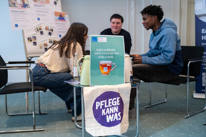 Drei Personen sitzen um einen Tisch in einem Raum. Auf dem Tisch steht ein Schild mit der Aufschrift "HAVE A BREAK, HAVE A CUP OF TEA FOR CARE" und "PFLEGE KANN WAS". Eine Person bietet eine Teekanne an, während die anderen beiden miteinander sprechen.