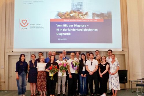 Eine Gruppe von 18 Personen steht im Vordergrund und hält Blumen. Im Hintergrund befindet sich eine Projektionsfläche mit dem Titel „Vom Bild zur Diagnose – KI in der kinder-kardiologischen Diagnostik“ und dem Datum 21. Juni 2025.