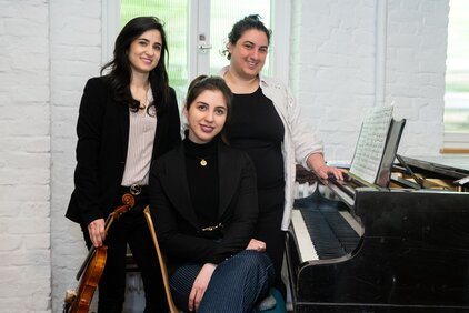 Drei Frauen stehen und sitzen in einem hellen Raum. Eine Frau hält eine Violine, während die anderen beiden lächeln, eine steht neben einem Klavier mit Notenblättern. Der Fokus liegt auf ihrer musikalischen Zusammenarbeit und Freundschaft.