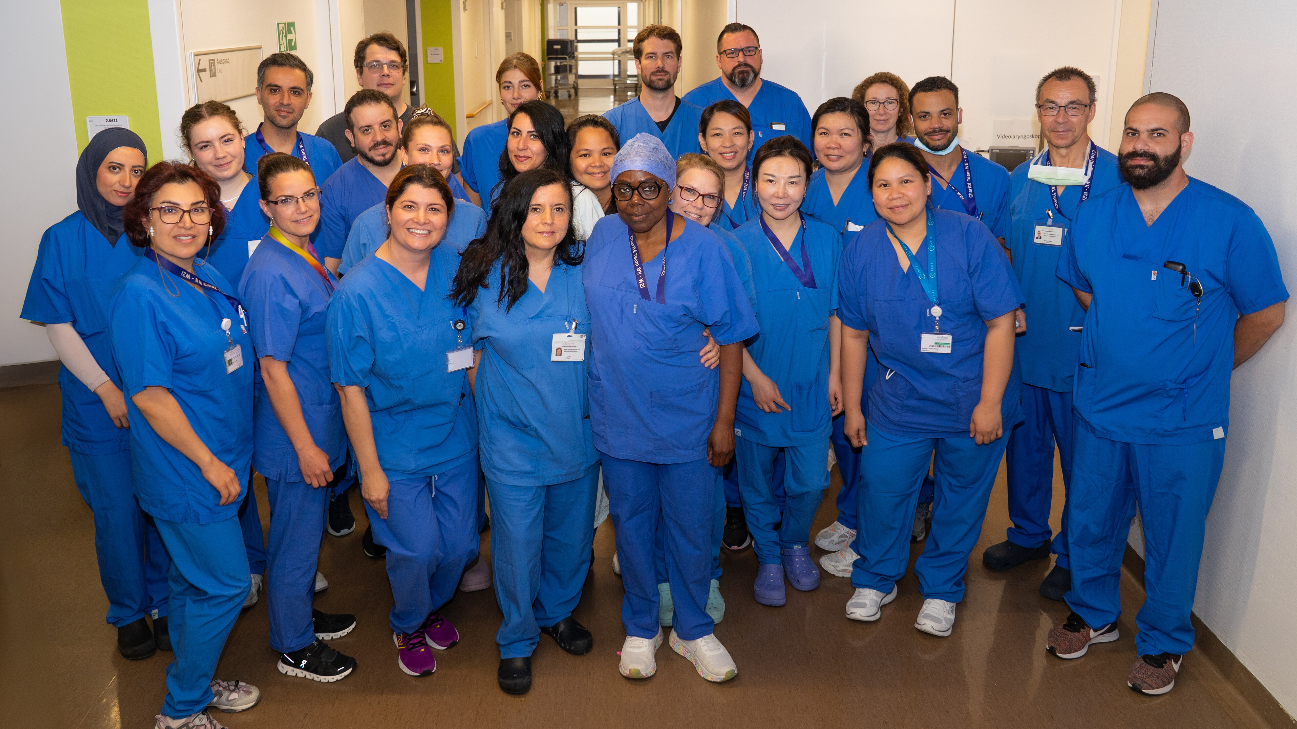 Gruppenszene von 24 Gesundheitsfachkräften in blauen Arbeitskleidungen, die vor einem Krankenhausflur posieren. Einige tragen Ausweise und Kopfbedeckungen. Die Atmosphäre wirkt freundlich und professionell. Im Hintergrund sind Wegweiser und eine Tür sichtbar.