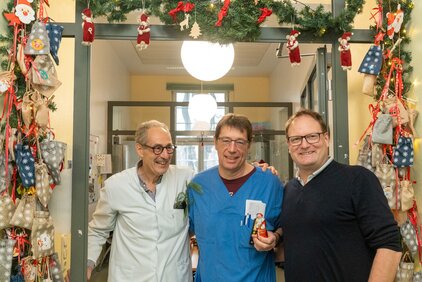 Drei Männer stehen in einem festlich dekorierten Eingangsbereich, der mit Weihnachtsdekoration geschmückt ist. Sie lächeln und posieren zusammen. Einer der Männer hält eine kleine Figur in der Hand, während ein anderer einen Zweig in der Tasche hat.