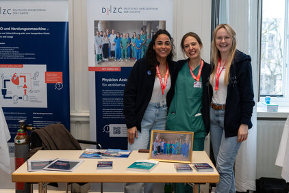 Drei Frauen stehen an einem Tisch, der mit Informationsmaterialien zum Deutschen Herzzentrum dekoriert ist. Jede trägt ein Namensschild und ein freundliches Lächeln. Im Hintergrund ist ein Banner mit Informationen zu Ausbildungsprogrammen sichtbar.