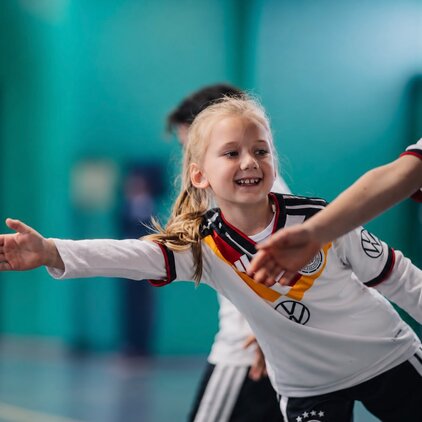Ein fröhliches Mädchen in einem Fußballtrikot mit Deutschland-Flagge spielt in einer Sporthalle. Sie lächelt und streckt ihren Arm aus, um mit einem anderen Kind zu interagieren. Die Umgebung ist grün, was zur sportlichen Atmosphäre beiträgt.