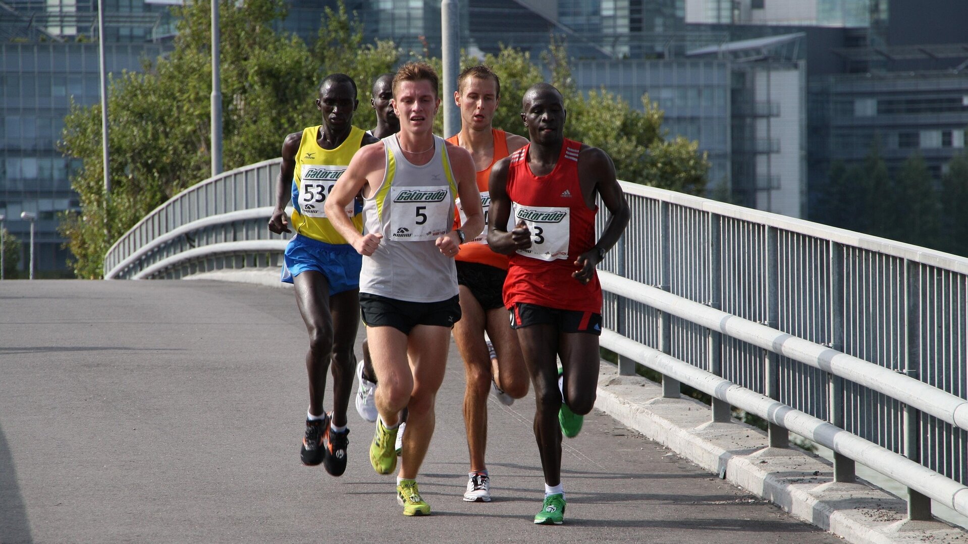 Vier Läufer nehmen an einem Wettkampf auf einer Brücke teil. Sie tragen farbige Sportbekleidung mit Nummern. Die Stimmung ist dynamisch und sportlich, während die Athleten konzentriert nebeneinander laufen. Die Umgebung besteht aus städtischen Gebäuden und Pflanzen.