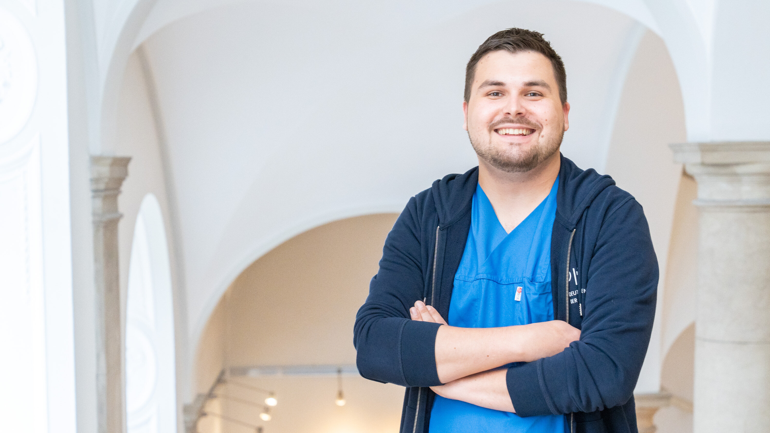Ein lächelnder Mann steht mit verschränkten Armen in blauer Arbeitskleidung. Er befindet sich in einem hellen, modernen Raum mit Bögen und Säulen im Hintergrund. Der Ausdruck auf seinem Gesicht wirkt freundlich und offen.