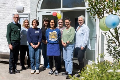 Gruppenbild von zehn Personen, die vor einem Gebäude stehen. Sie lächeln und tragen legere Kleidung. Im Hintergrund sind bunte Luftballons und Pflanzen sichtbar. Das Bild vermittelt eine freundliche und einladende Atmosphäre.