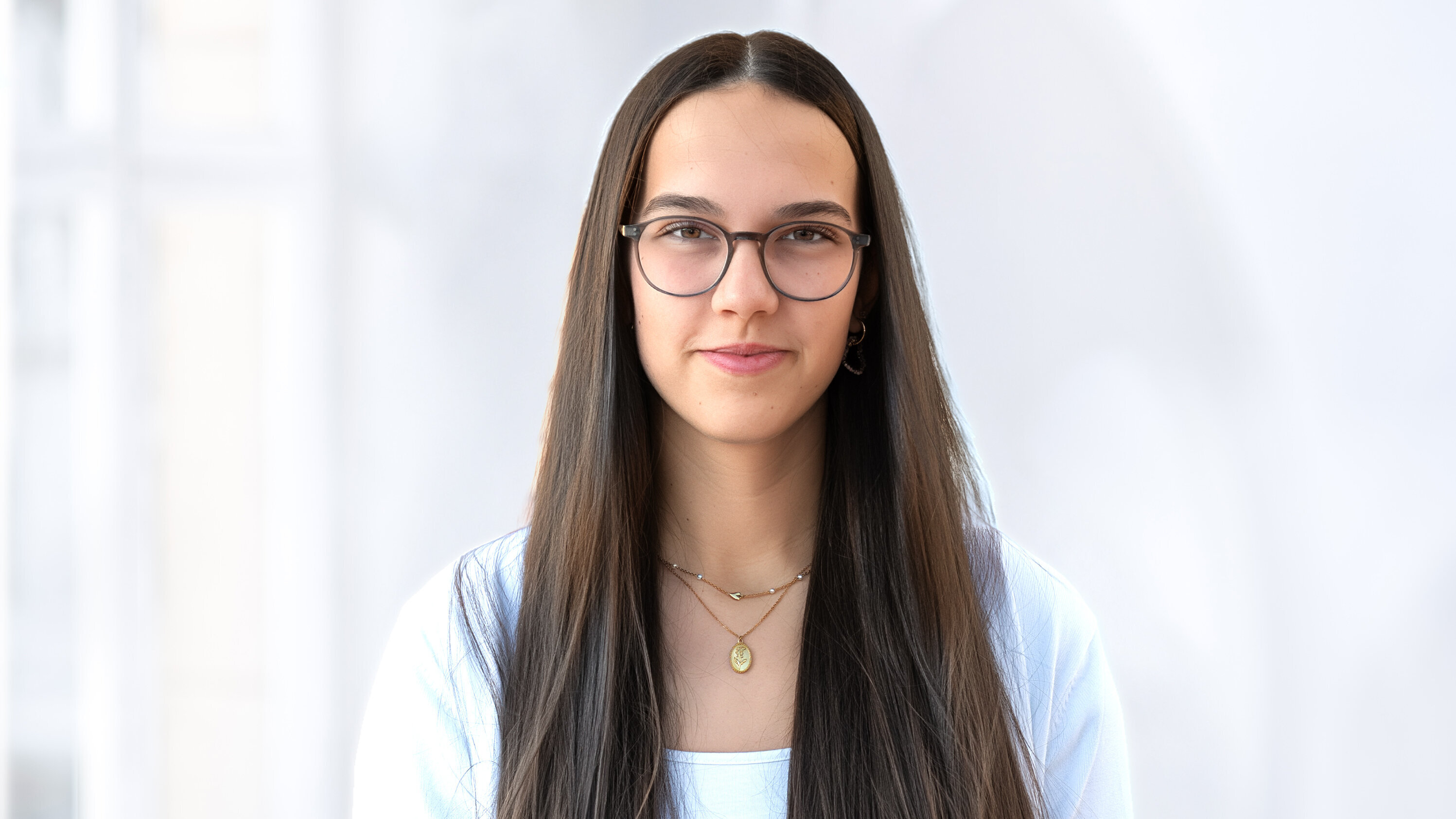 Eine junge Frau mit langen, dunklen Haaren steht vor einem hellen Hintergrund. Sie trägt eine weiße Bluse und eine goldene Halskette. Ihre Brille hat einen zarten, modischen Rahmen. Ihr Ausdruck ist freundlich und selbstbewusst.